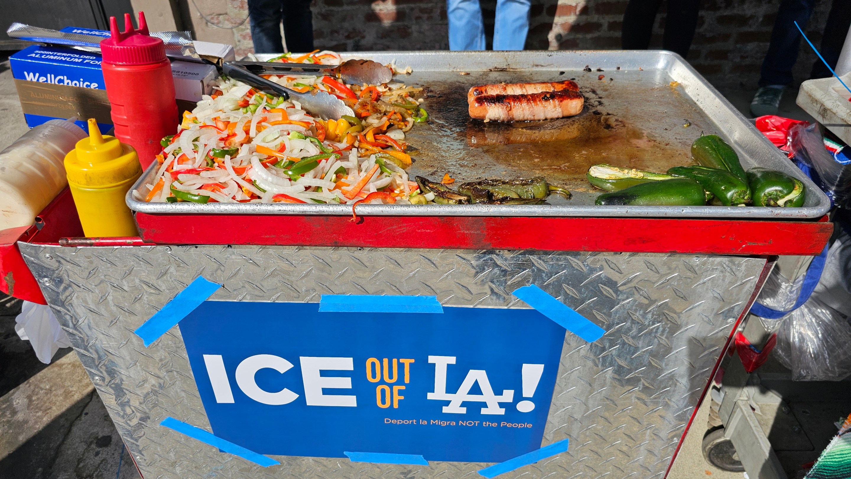 A street vending cart topped with bacon wrapped hotdogs and grilled veggies. There is an "ICE out of LA!" poster on the front of the cart.