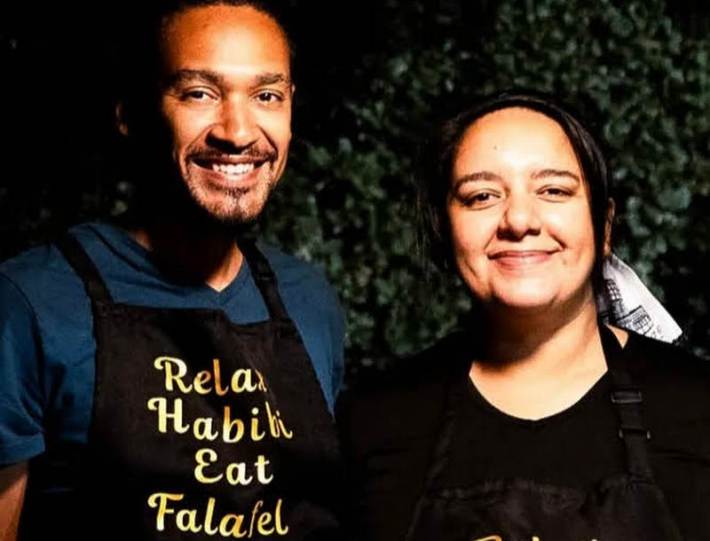 A couple together, he wears a black apron that says "Relax Habibi Eat Falafel"