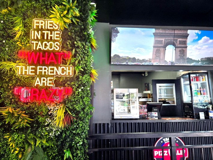 Neon sign on a background of green vines that says "Fries in the tacos? The French are crazy."