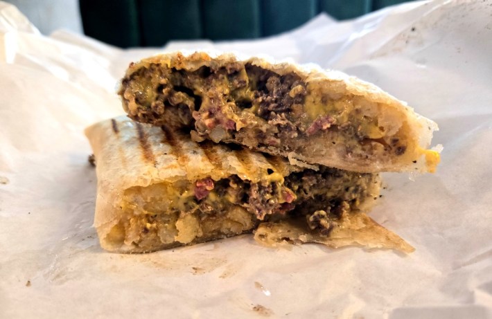 A French tacos, sliced in half showing beef, beef bacon, fries, and cheese in a flour tortilla.