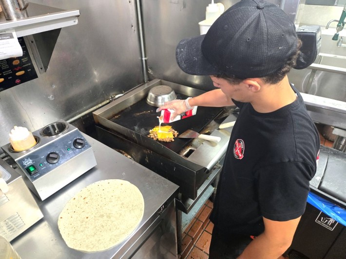 A man in a black t--shirt cooks over a griddle, with a big flour tortilla at his side