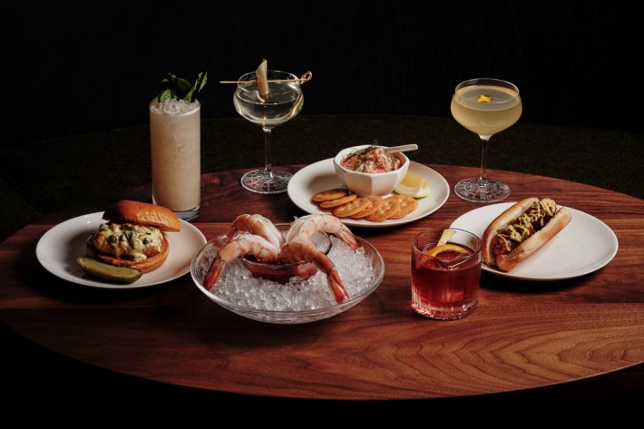 A table set with a hot dog, fish sandwich, giant shrimp in a bowl full of ice, and a crab dip with crackers, surrounded by various cocktails