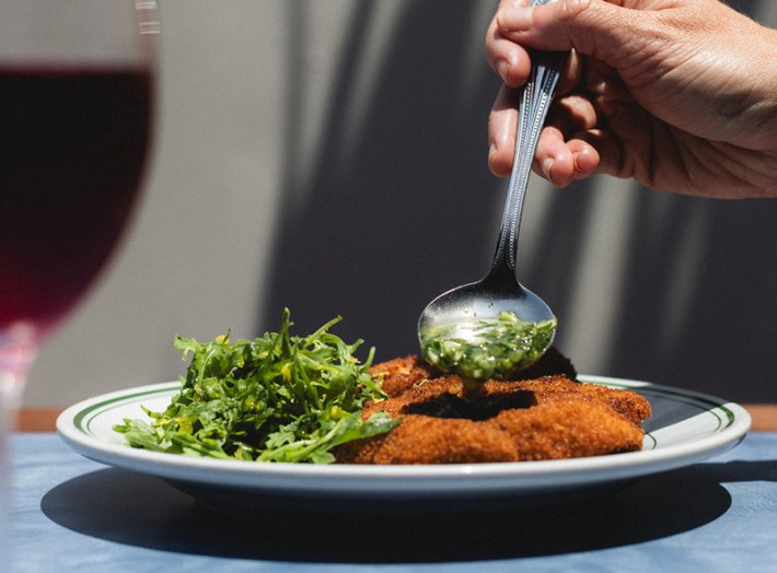 A hand holds a spoon, drizzling chimichurri onto a chicken milanesa, with a salad on the side