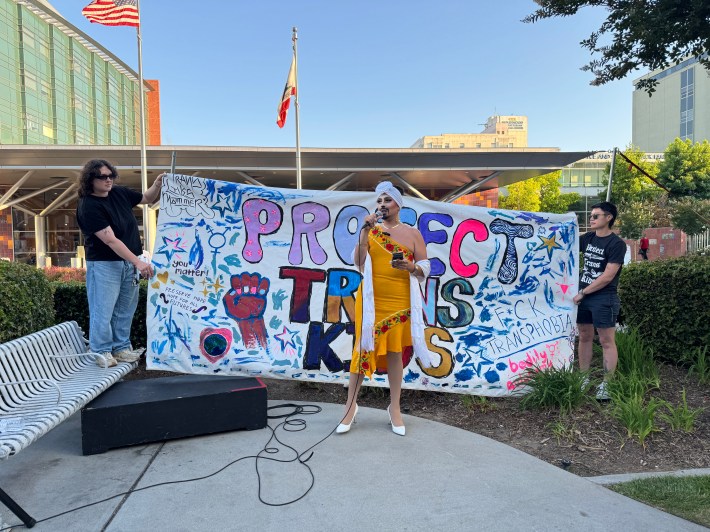 Drag artist ChiChi Charlas speaks in front of CHLA, in a yellow dress and turban, while holding a microphone.