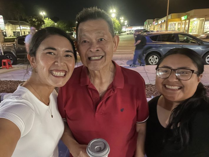 A smiling woman stands next to an older man in a red shirt and a younger, smiling woman at a night market in L.A.