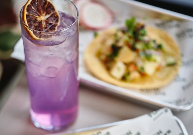 A purple mocktail with a dehydrated orange garnish and a blurry tostada in the back