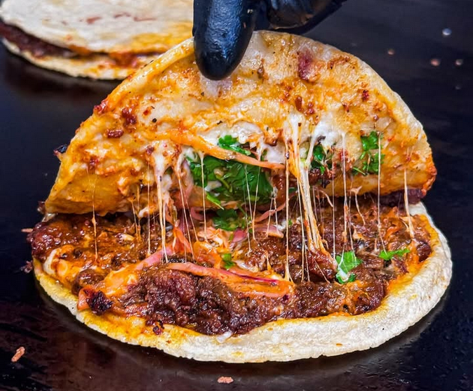 A black gloved hand pulls back a mulita, revealing meat and cheese. Photo via Balam.