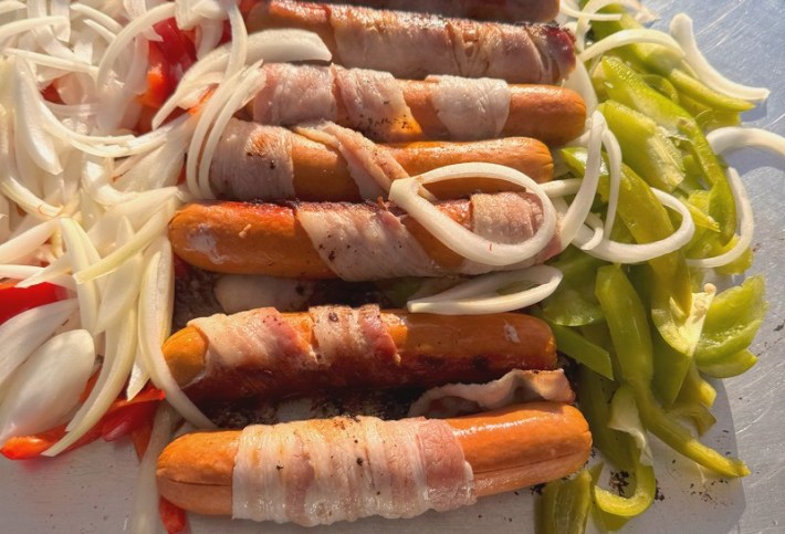Seven bacon-wrapped hot dogs grilling with onions and peppers