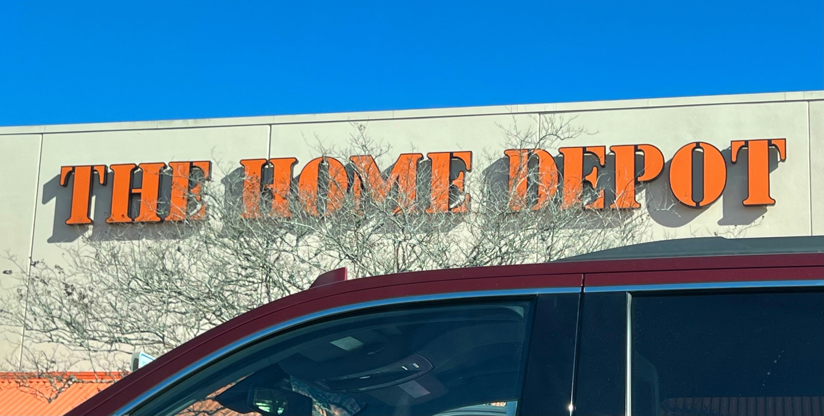 A Home Depot sign and roof in front of a blue sky, with car windows passing below