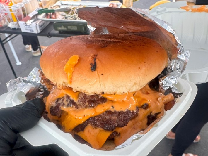 A double smashburger with cheese in a styrofoam box, held by a black gloved hand