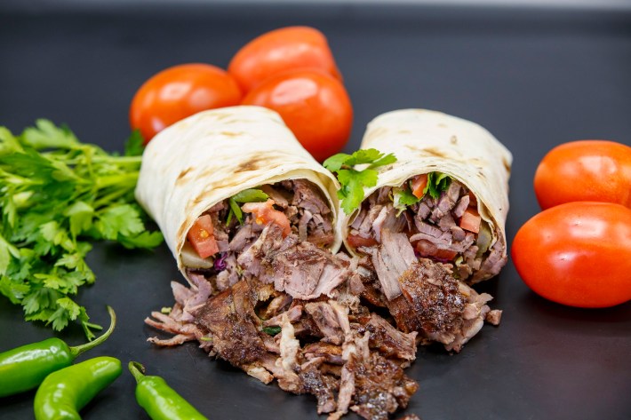 A split doner wrap spilling meat on the table next to loose cilantro, chiles, and tomatoes