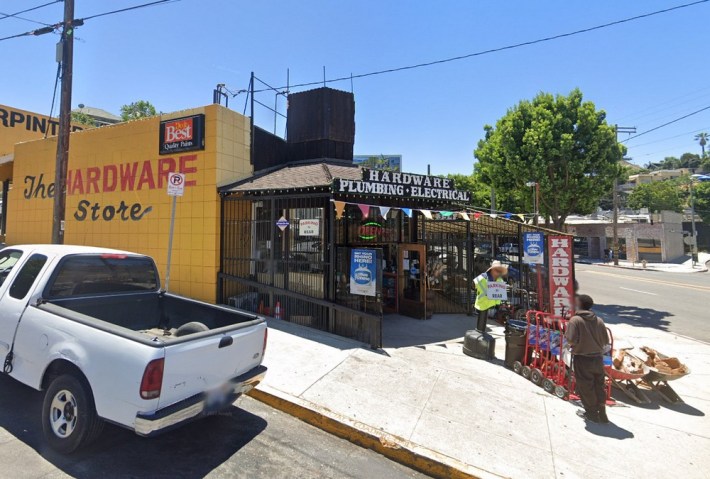 A curbside hardware store with an employee holding a sign and a customer approaching, a white truck parked on the side