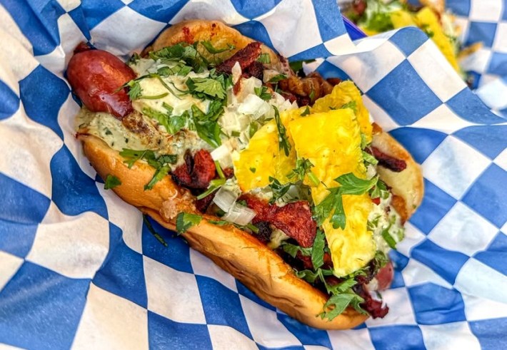 A pineapple-and-pastor-loaded hot dog with cilantro and cilantro crema, on a background of blue checke