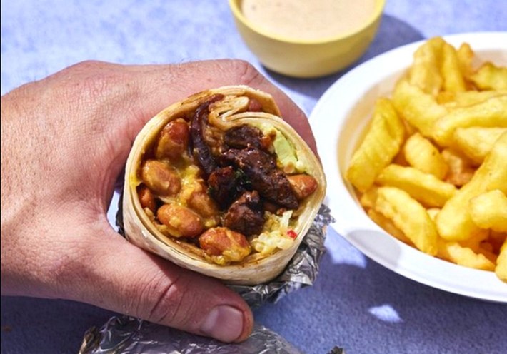 A hand holds a burrito half filled with beans and beef, next to a plate of fries