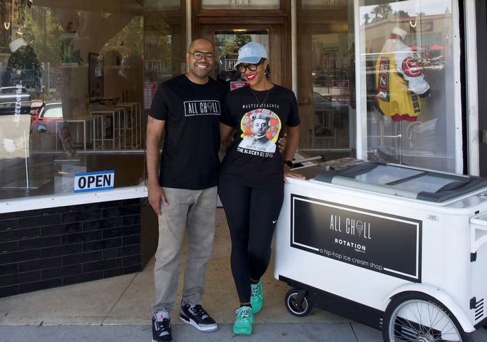 A couple stands in front of their ice cream shop, with an ice cream cart at their side