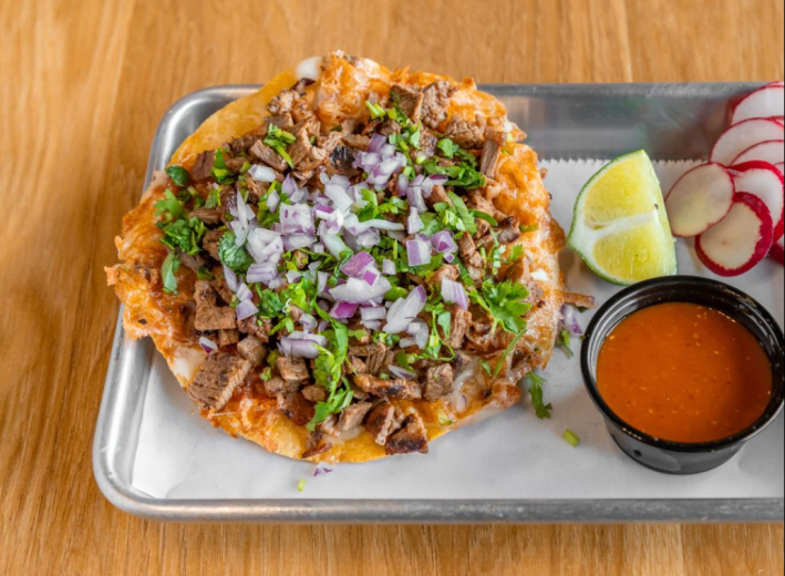A Vampiro with birria, cilantro, and chopped red onions, on a tray next to a cup of salsa roja, radishes, and a wedge of lime