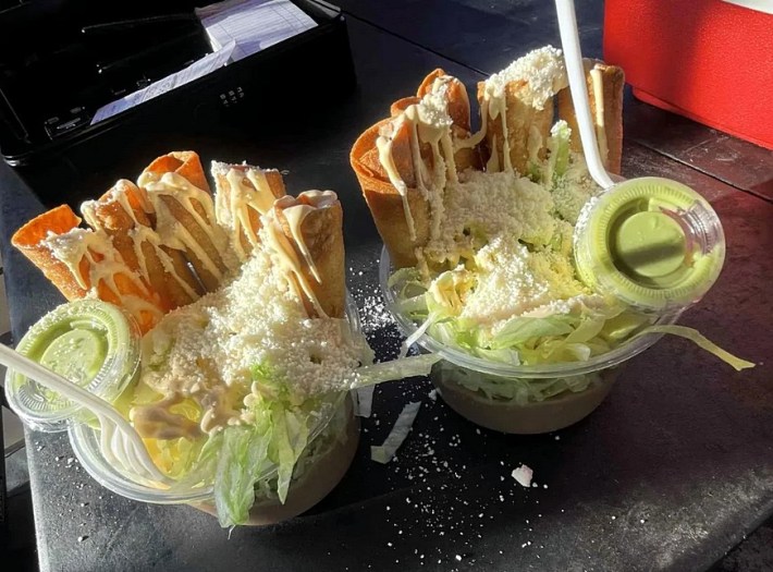Two plastic cups full of taquitos filled with shredded lettuce and topped with cotija cheese, each with a tiny cup of guacamole