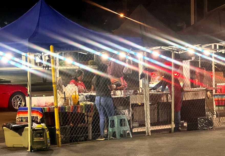 A view of a taco stnd in Altadena from the back, showing a fence, lighting, and taqueras