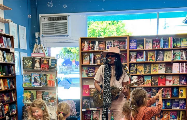 A man with a guitar and broad brimmed hat sings to a group of children at a bookstore