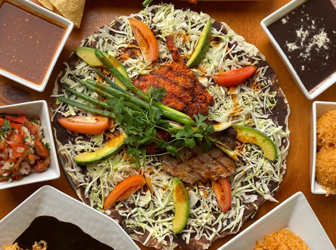 A tlayuda with cheese, beans, asiento, tomato, avocado, crhoizo and asada, ringed with bowls of salsas and sides like beans and rice