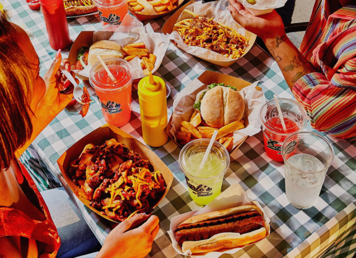 Hot dogs, lemonade, punch, and chili at a table with a black and white checkerboard tablecloth, with two people barley peeking into the photos