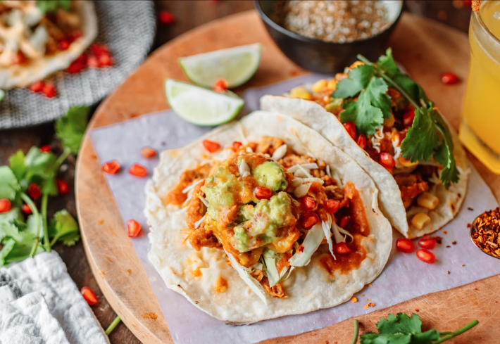 Two tacos with orange sauce and pomegranate seeds, with cilnatro and limes blurry in the background