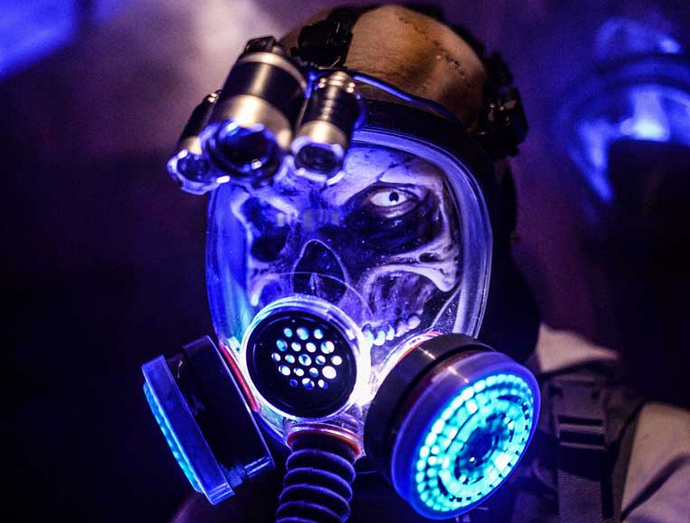 A grinning skull behind a gas mask and faceshield in blue light