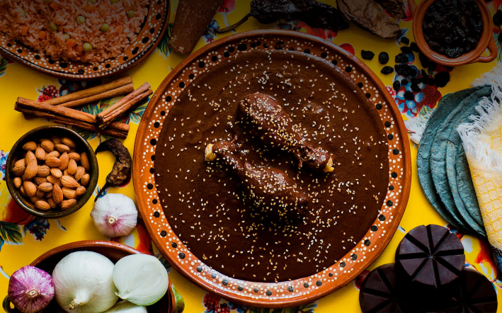 Two chicken legs in a big bowl of mole sprinkled with sesame seeds and surrounded by cinnamon, garlic, and almonds