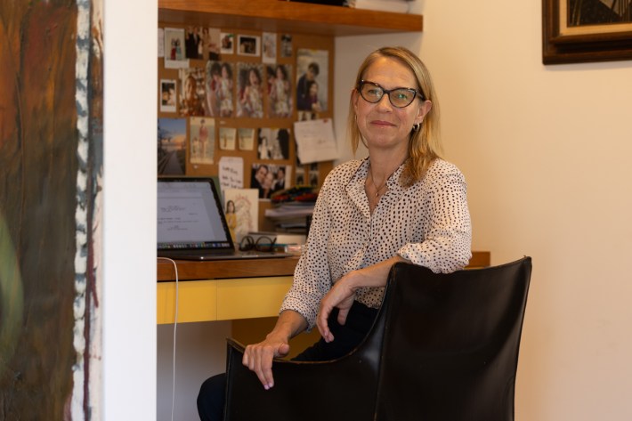 A blonde woman in glasses sits at her desk in her Los Angeles home, where she normally writes, in front of a laptop and photos pinned to a board
