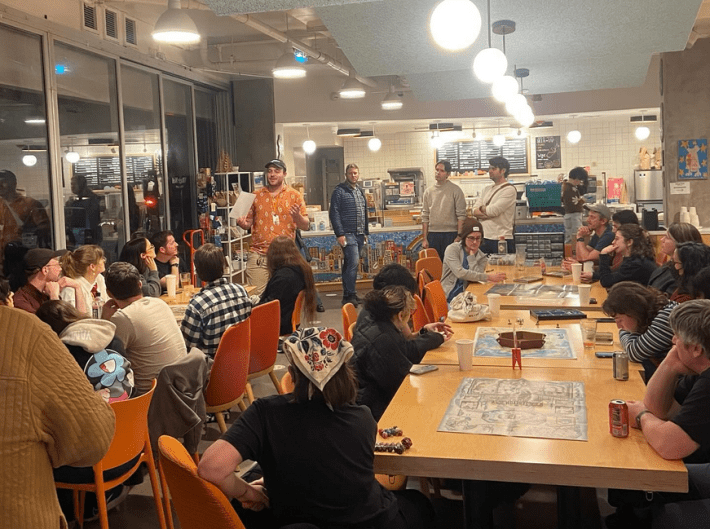 A man in a red shirt talks to a gathering of people at cafe tables.