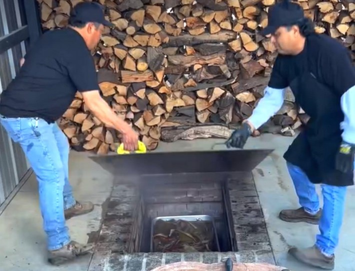 Two men open a pit in the floor for roasting barbacoa, with smoke being released and a background of stacked wood