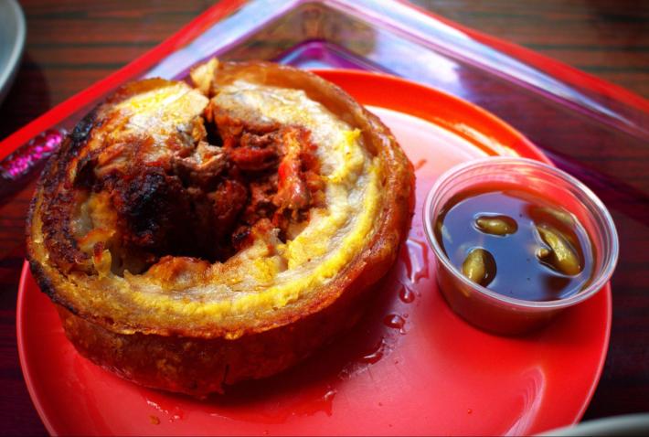 A thick circular cut of chicharron next to a small dish of an oil-based sauce, on a large red plate