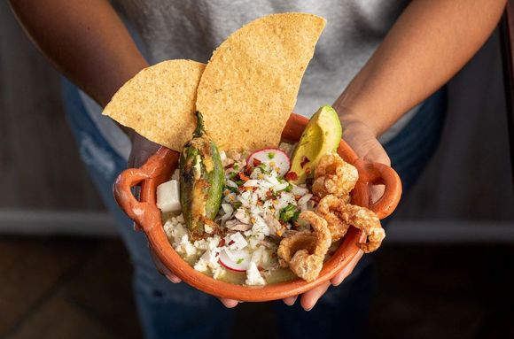 A bowl of pozole blanco in an orang ceramic pot, with tostadas and chicharron on top, in a woman's hands