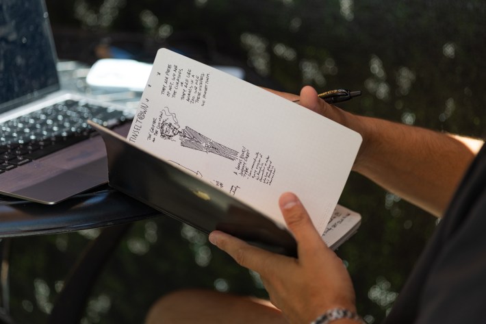 A man flips through his notebook while working at a coffee shop, showing a sketch of a man in a suit, smoking