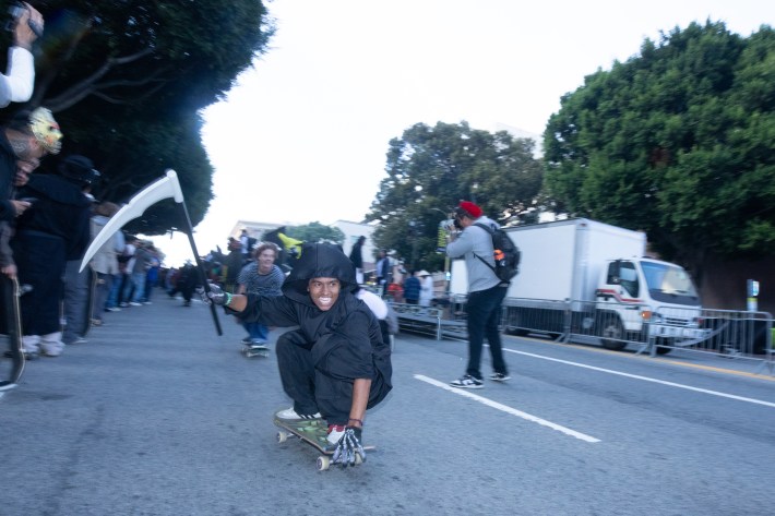 someone skates down a street wearing a black cloak and holding a fake scythe
