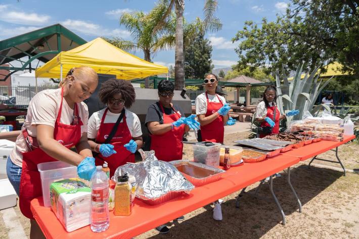 Women wearing aprons stand behind a table with food platters set up.