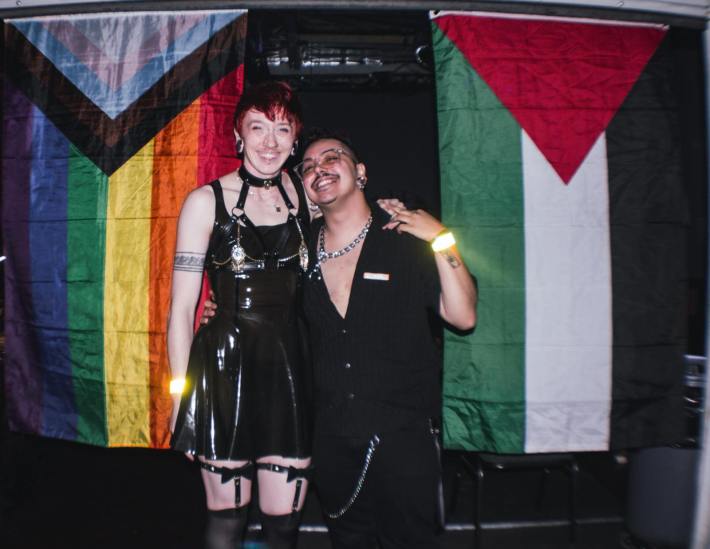 A couple, one wearing a black latex dress with short red hair and leather harness and one with facial hair wearing all black, smile together in front of a pride flag and Palestinian flag.