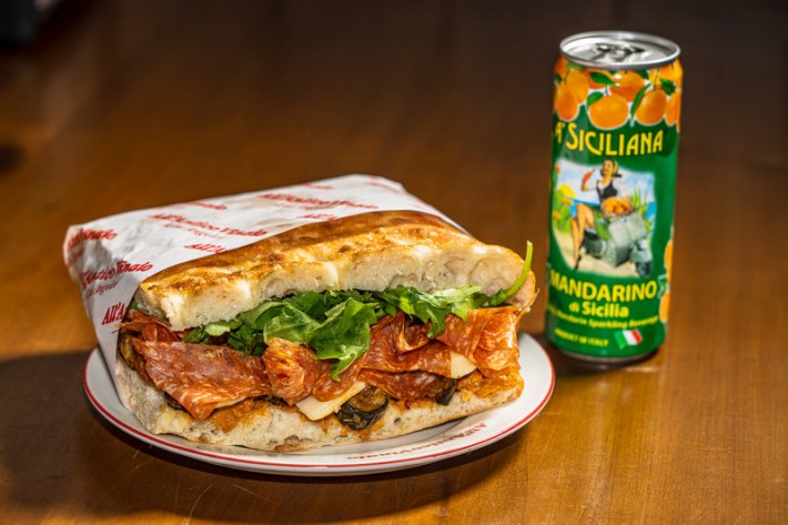 A paper-wrapped sandwich filled with salami and spinach next to a green can of Italian soda