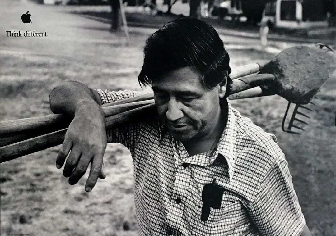 Cesar Chavez looks down while holding farming tools on his shoulder.