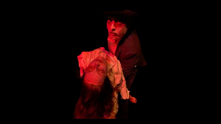 A man with a beard and dark clothing holds a woman's lifeless body, titling her towards the camera