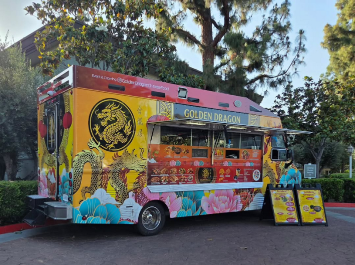A yellow and red Chinese food truck named Golden Dragon, parked with its sandwich boards