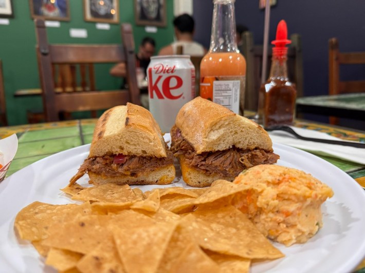 A split cochinita pibil torta in front of a bottle of hot sauce and a can of Diet Coke, with chips in the foreground