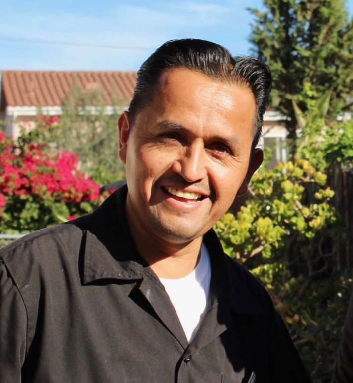 A man in a collared shirt smiles in front of flowers and bushes.