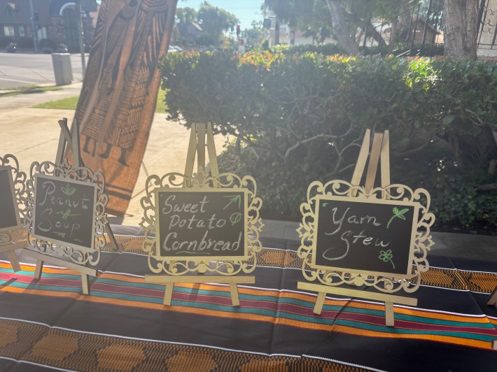 Three little signs are set up on a table: "Peanut Soup," "Sweet Potato Cornbread," and "Yam Stew."