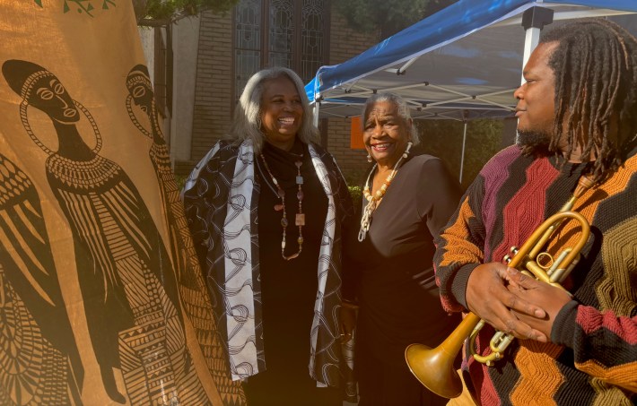 Two women and a man holding a trumpet pose next to a tapestry showing African artwork.