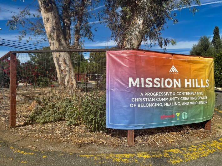 A large rainbow-colored banner says "MISSION HILLS" and describes the church's goals beneath it. This is placed on the fence of a garden.