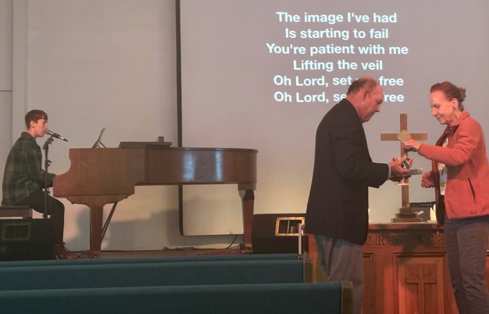 A woman sings and plays the piano while a man and woman exchange communion.