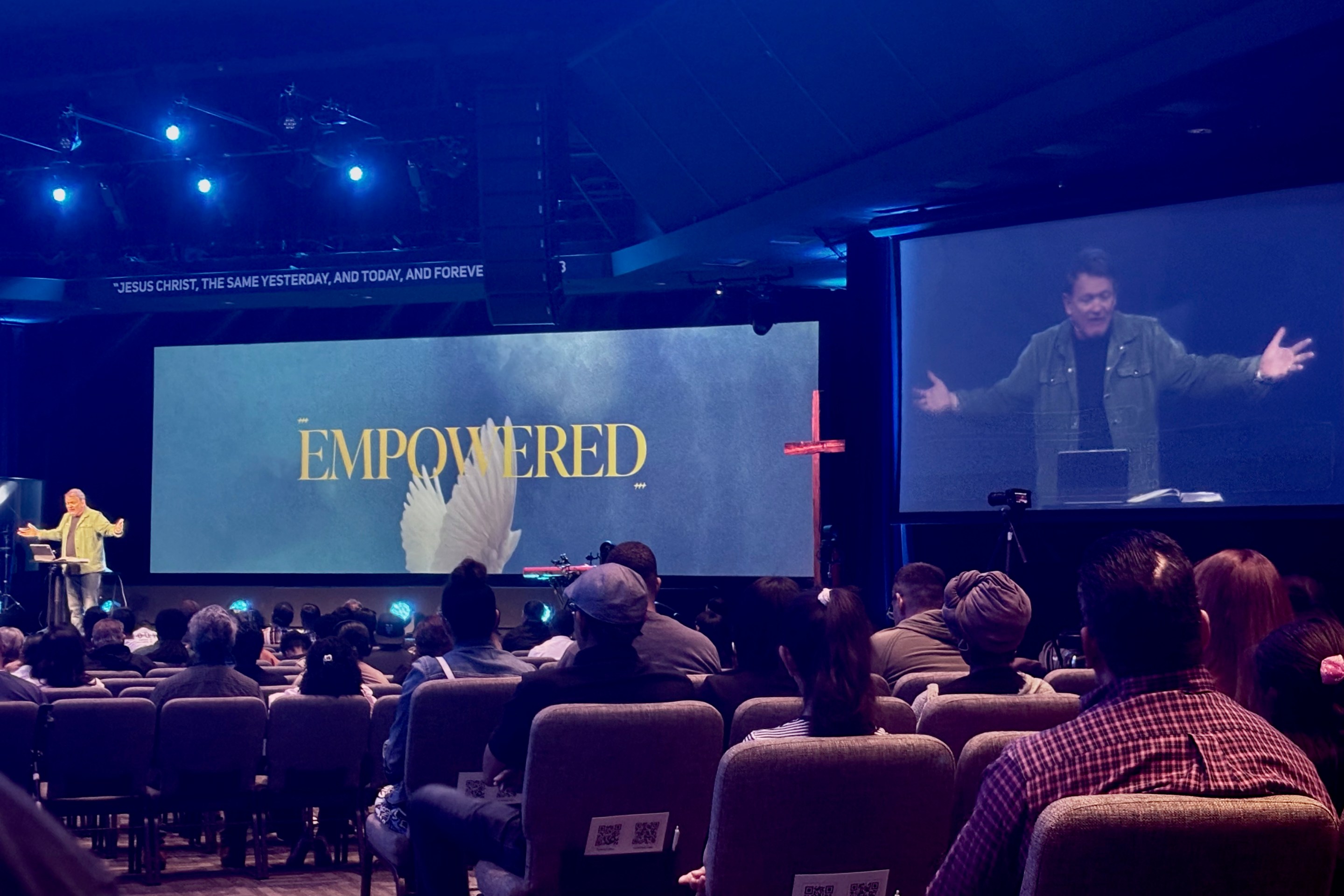 A man preaches from a stage in front of a crowd. A large digital screen is behind him with the word "empowered" on it.