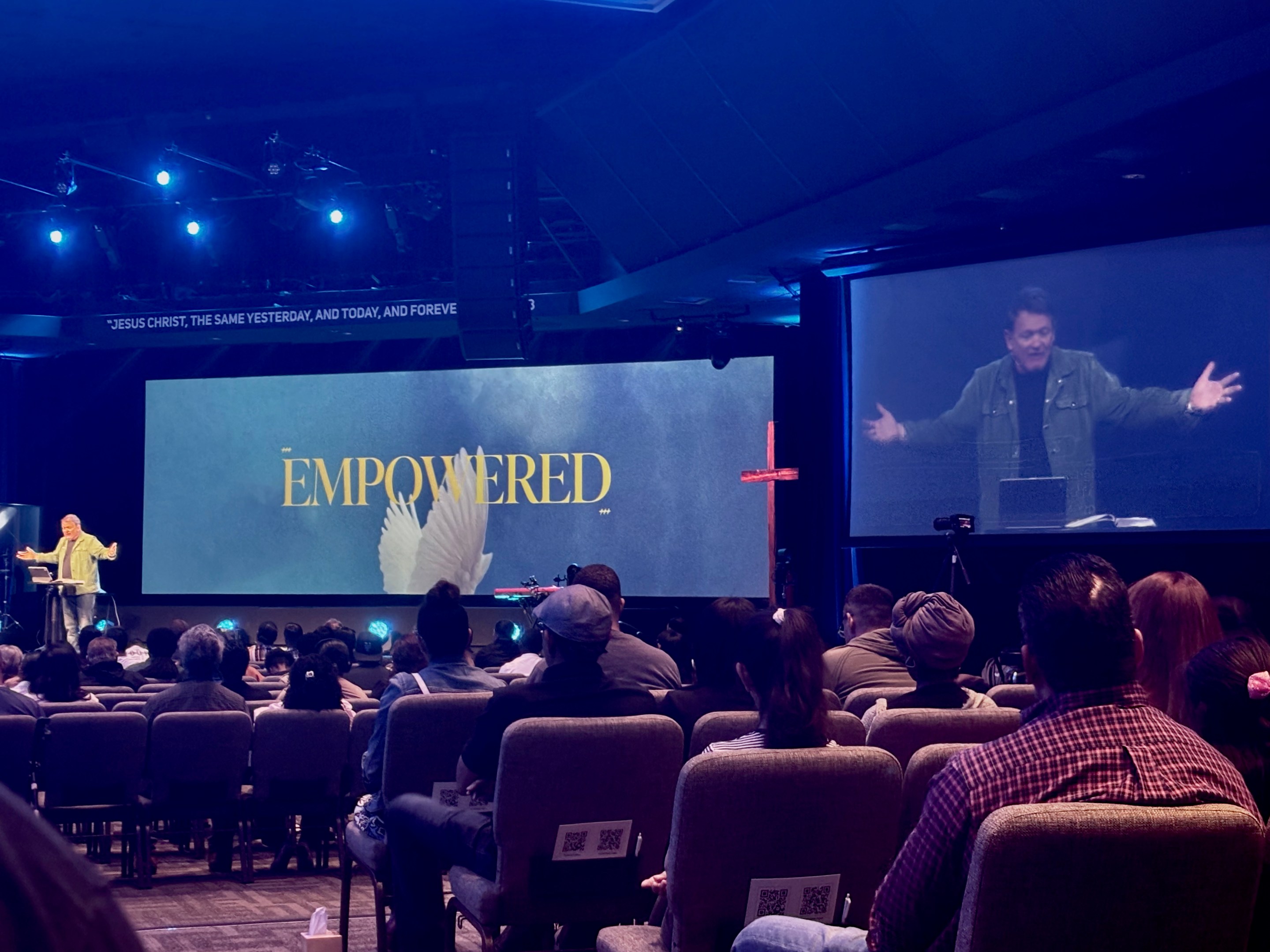 A man preaches from a stage in front of a crowd. A large digital screen is behind him with the word "empowered" on it.