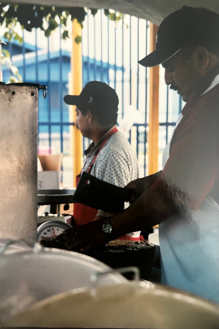A man in a baseball cap cuts a slab of meat on a tree trunk slab.